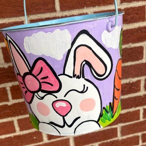 May include: A hand-painted Easter bucket with a light purple exterior, featuring a smiling white bunny with a pink bow, pink cheeks, and a carrot. The bucket has a light blue handle and rim.
