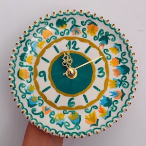 May include: A round wall clock with a white ceramic base featuring a hand-painted floral pattern in teal, yellow, and orange. The clock face has a dark teal center, gold-colored hands, and numbers 3, 6, 9, and 12 painted in a dark teal.
