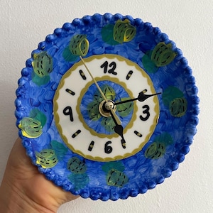 May include: A round ceramic wall clock featuring a hand-painted blue background with green and yellow abstract patterns. The center has a white dial with black numerals 3, 6, 9, and 12, surrounded by a gold-toned border and black clock hands.