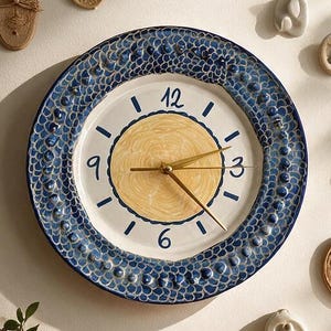 Könnte beinhalten: Eine runde Wanduhr aus Keramik mit einem gelben Mittelkreis, einem weißen Zifferblatt mit blauen Ziffern 3, 6, 9 und 12 sowie einem strukturierten blauen Rand mit erhabenen Punkten. Die Uhr hat dünne goldfarbene Zeiger und ist von kleinen Wandobjekten umgeben.