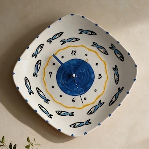 May include: A square-shaped wall clock with rounded corners, featuring a white ceramic-style face decorated with a pattern of small blue and black fish. The center has a dark blue circular design with black numbers 3, 6, 9, and 12.