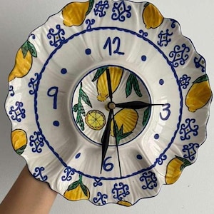 May include: A round ceramic wall clock with a scalloped edge, featuring a hand-painted pattern of yellow lemons and blue decorative motifs on a white background. The clock face displays blue numerals at 12, 3, 6, and 9, with three black hands.