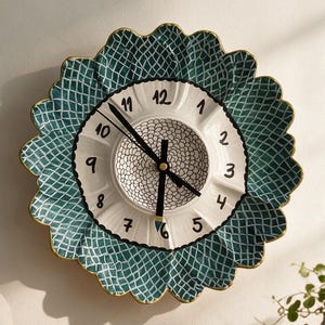 Puede incluir: Un reloj de pared redondo con borde festoneado que presenta un patrón de cuadrícula en verde azulado oscuro y blanco. El centro es blanco con números negros del 1 al 12 y un detalle circular texturizado. Tiene tres manecillas negras.
