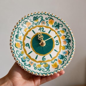 May include: A round ceramic wall clock with a scalloped edge, featuring a hand-painted floral pattern in yellow, blue, and green. The clock face has a dark green center, gold-painted numerals, and gold-toned metal hands against a white background.
