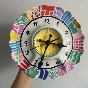 May include: A colorful, hand-painted ceramic clock with a white face and black numbers. The clock has a scalloped edge and is decorated with abstract shapes in various colors, including red, yellow, blue, green, and pink.