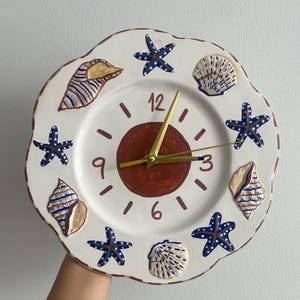 May include: A white ceramic clock with a red center and gold hands. The clock is decorated with blue and brown seashells and starfish. The numbers on the clock face are 12, 9, 6, and 3.