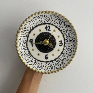 Puede incluir: Un reloj de pared redondo con un borde festoneado pintado en dorado. El borde exterior presenta un patrón negro dibujado a mano sobre un fondo blanco. El centro tiene un círculo negro con puntos blancos, marcadores horarios dorados y los números negros 3, 6, 9 y 12.
