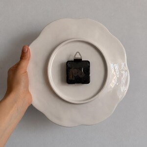K&ouml;nnte beinhalten: Die R&uuml;ckseite einer wei&szlig;en Wanduhr aus Keramik mit gewelltem Rand. Ein schwarzes, batteriebetriebenes Uhrwerk aus Kunststoff ist in der Mitte montiert und verf&uuml;gt &uuml;ber einen kleinen Metallaufh&auml;nger an der Oberseite zur Wandmontage.