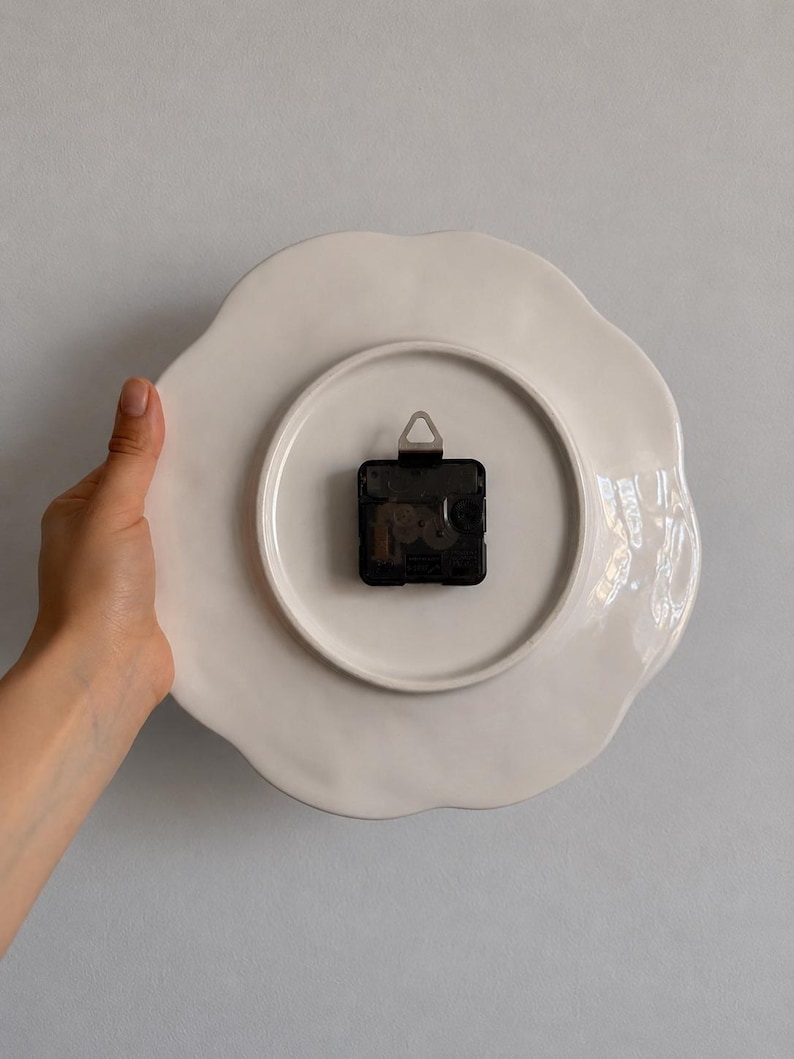 K&ouml;nnte beinhalten: Die R&uuml;ckseite einer wei&szlig;en Wanduhr aus Keramik mit gewelltem Rand. Ein schwarzes, batteriebetriebenes Uhrwerk aus Kunststoff ist in der Mitte montiert und verf&uuml;gt &uuml;ber einen kleinen Metallaufh&auml;nger an der Oberseite zur Wandmontage.