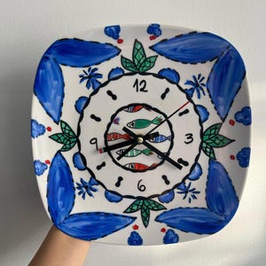 May include: A square ceramic wall clock with a white background, featuring hand-painted blue leaf motifs, green accents, and a central circular pattern of small red, green, and blue fish. The clock face displays black numbers 3, 6, 9, and 12.