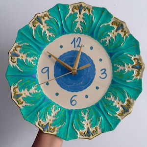 May include: A round wall clock featuring a scalloped edge with a vibrant turquoise and gold leaf-like pattern. The center has a cream-colored face with a blue circular accent, blue numerals 12, 3, 6, and 9, and gold-toned clock hands.