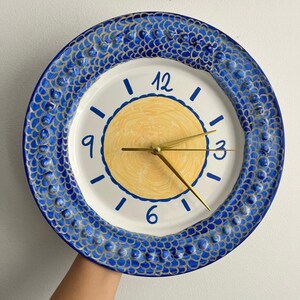 May include: A round wall clock featuring a white face with a central yellow circle, blue numerals at 3, 6, 9, and 12, and gold-colored hands. The outer rim is decorated with a textured, blue and white scale pattern with raised circular details.