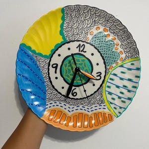 May include: A decorative wall clock made from a plate, featuring a colorful, hand-painted design. The clock face is white with black numerals and hands. The plate's rim is decorated with yellow, blue, and orange sections, and black and white patterns.