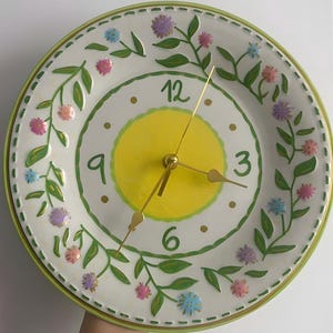 Puede incluir: Un reloj de pared redondo con esfera blanca y un círculo amarillo central. El reloj tiene los números 3, 6, 9 y 12 en verde y manecillas doradas. El borde está decorado con enredaderas verdes pintadas a mano y pequeñas flores rosas, moradas y azules.