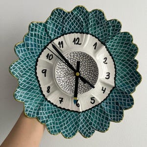 Può includere: Un orologio da parete decorativo in ceramica con un design unico a forma di girasole. L'orologio presenta un quadrante bianco con numeri e lancette nere. Il bordo esterno è a forma di conchiglia con un motivo turchese e bianco. L'orologio misura circa 25 cm di diametro.