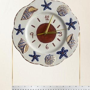 K&ouml;nnte beinhalten: Eine runde, handbemalte Keramik-Wanduhr mit wei&szlig;em Grund, einem dunkelroten Mittelkreis, goldfarbenen Zeigern und den Ziffern 3, 6, 9 und 12. Der Rand ist mit blauen Seesternen und beigen Muscheln verziert. Der Durchmesser betr&auml;gt 26,67 cm.