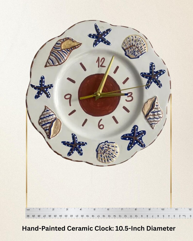 K&ouml;nnte beinhalten: Eine runde, handbemalte Keramik-Wanduhr mit wei&szlig;em Grund, einem dunkelroten Mittelkreis, goldfarbenen Zeigern und den Ziffern 3, 6, 9 und 12. Der Rand ist mit blauen Seesternen und beigen Muscheln verziert. Der Durchmesser betr&auml;gt 26,67 cm.