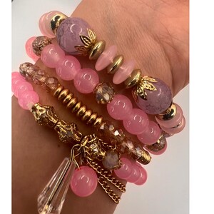 Könnte beinhalten: Ein Set aus fünf rosafarbenen und goldenen Armbändern mit verschiedenen Perlenstilen und einem goldenen Anhänger. Die Armbänder haben rosafarbene und violette Perlen, goldene Perlen und eine goldene Kette mit einem rosafarbenen Anhänger.