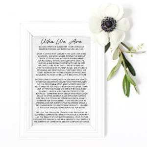 May include: A framed print with the text "Who We Are" and a description of a mother-daughter team. A white anemone flower and green leaves are in the upper right corner. The frame is white.