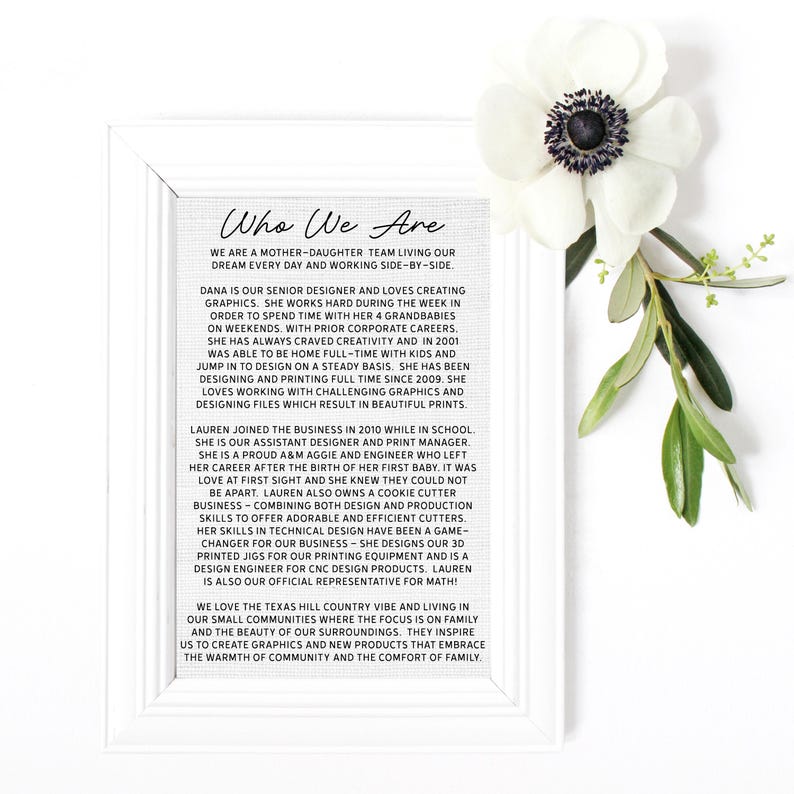 May include: A framed print with the text "Who We Are" and a description of a mother-daughter team. A white anemone flower and green leaves are in the upper right corner. The frame is white.