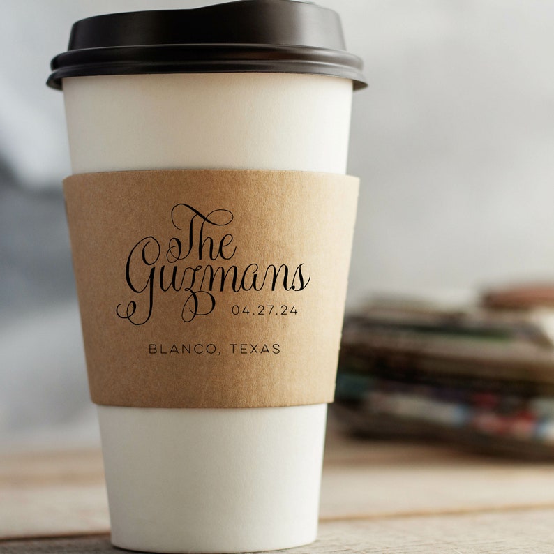 May include: A white paper coffee cup with a brown cardboard sleeve. The sleeve features the text "The Guzmans" above the date "04.27.24" and the location "BLANCO, TEXAS" in black script. The cup has a black lid.