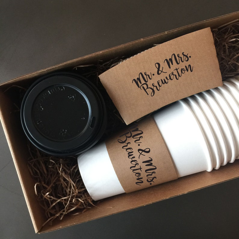 May include: A gift box containing white paper coffee cups, a black lid, and brown cardboard sleeves. The sleeves and one cup have the text "Mr. & Mrs. Brewerton" in black script. The box is filled with brown packing material.