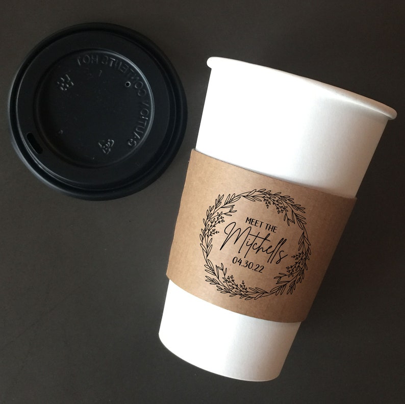 May include: A white paper coffee cup with a brown paper sleeve featuring a floral wreath design and the text "Meet the Mitchells 04.30.22." A black plastic lid sits next to the cup. The cup is on a dark surface.