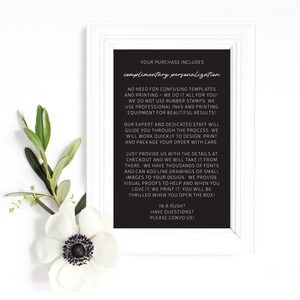 May include: A framed black sign with white text, including "complimentary personalization." The sign is surrounded by a white frame and decorated with a white flower and green leaves. The text describes the personalization process.