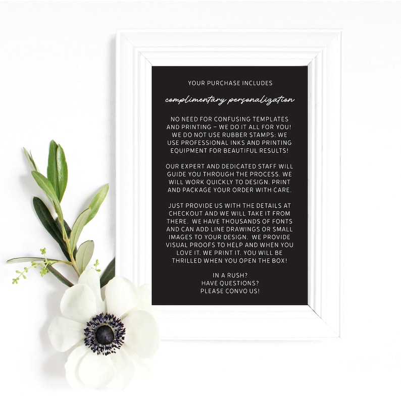 May include: A framed black sign with white text, including "complimentary personalization." The sign is surrounded by a white frame and decorated with a white flower and green leaves. The text describes the personalization process.