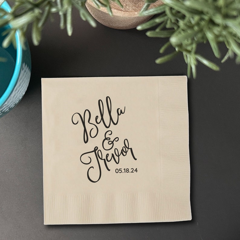 May include: A beige paper napkin with black calligraphy that reads "Bella & Trevor" and the date "05.18.24".