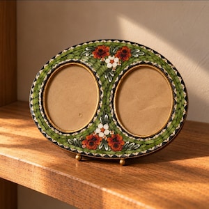 May include: An oval double photo frame featuring a mosaic design with green, white, and orange-red floral patterns. The frame has two circular openings with gold-toned rims and is supported by two small gold-toned metal feet at the base.