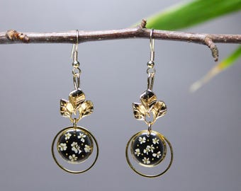 Puede incluir: Pendientes de oro con un colgante circular negro con detalles de flores blancas. El colgante está rodeado por un anillo dorado. Los pendientes tienen un diseño de hojas por encima del colgante.