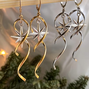 May include: A pair of gold and silver dangle earrings with a star design. The earrings are twisted and have a long, flowing shape.