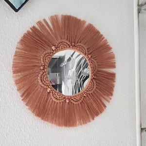 May include: Round wall mirror with a macrame design in shades of brown. The mirror has a fringe of long, thin strands radiating outwards. The inner circle is a woven pattern with small decorative beads. The mirror is hanging on a white wall.