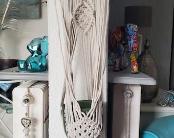 May include: A beige macrame plant hanger with a wooden dowel at the top. The woven design features a diamond pattern and a fringe at the bottom. A green pot is visible inside the hanger. Decorative items are in the background.