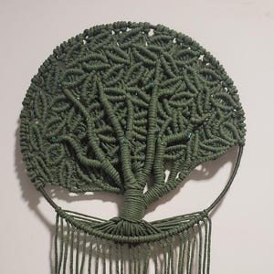 Può includere: Applique da parete in macramè verde oliva con design ad albero della vita. Il pezzo circolare presenta un motivo di annodatura dettagliato per le foglie e un tronco centrale con rami. Lunghe frange annodate pendono dal basso, aggiungendo consistenza e dimensione.