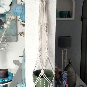 May include: A macrame plant hanger with a green ceramic pot. The off-white rope hanger is knotted and woven, holding the pot. The pot has a white dotted design. The overall theme is home decor.