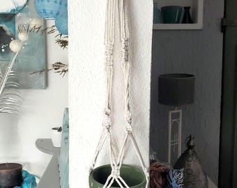 May include: A macrame plant hanger with a green ceramic pot. The off-white rope hanger is knotted and woven, holding the pot. The pot has a white dotted design. The overall theme is home decor.