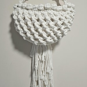 Peut inclure: Un panier en macramé fixé au mur, fabriqué à partir de corde de coton blanc cassé. Le panier présente un corps tissé semi-circulaire avec un motif en treillis noué, une poignée supérieure incurvée pour la suspension et une longue frange épaisse.