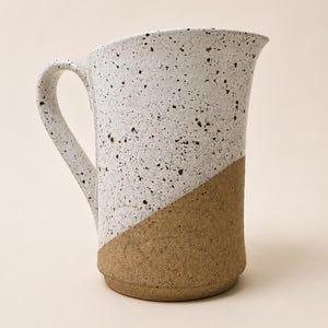 May include: A ceramic pitcher featuring a two-tone design with a speckled white upper section and a solid, textured tan base. The vessel includes a curved handle on the left side and a wide, angled pouring spout at the top rim of the container.