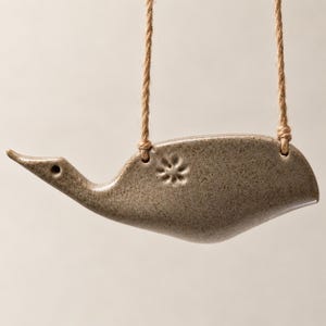 Peut inclure: Un ornement d'oiseau en céramique brun clair moucheté avec une finition lisse et brillante. L'oiseau présente un petit motif de fleur estampé sur le côté et est suspendu par deux fines ficelles de jute brun clair passant par des trous.
