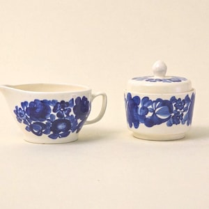 Könnte beinhalten: Ein cremefarbenes Sahne- und Zuckerdosen-Set. Beide Teile sind mit einem leuchtend blauen Blumenmuster verziert. Der Sahneaufgießer hat einen Griff und einen Ausguss, während die Zuckerdose einen Deckel mit einem kleinen Knopf hat.