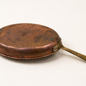 Könnte beinhalten: Eine runde Kupferpfanne mit einer verwitterten, dunkelbraunen Patina und einer strukturierten Oberfläche. Die Pfanne hat einen langen, flachen Metallgriff, der mit einer Nietverbindung befestigt ist. Das Metall zeigt Oxidationsspuren und Alter.