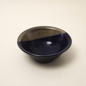 May include: A small, round ceramic bowl featuring a two-tone glaze. The interior and lower exterior are finished in a deep, glossy navy blue, while the upper rim area is coated in a contrasting matte, earthy brown tone with a smooth texture.