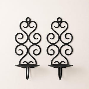 Puede incluir: Un par de candelabros de pared en negro mate con diseños de volutas simétricas. Cada pieza consta de una base circular para una vela, sostenida por un marco de metal curvado con un lazo integrado en la parte superior para colgar.