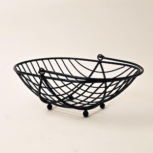 Puede incluir: Cesta de alambre de metal negro con forma ovalada. La cesta tiene un asa en cada extremo y se apoya en tres pequeños pies de bola. El diseño de alambre crea un patrón de celosía en toda la cesta.