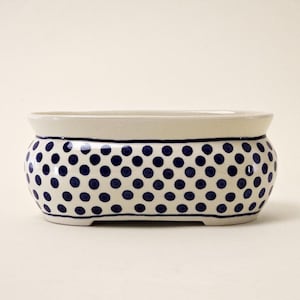 Peut inclure: Un pot de fleurs en céramique avec un bord et une base crème, et un corps recouvert de pois bleu marine. Le pot rectangulaire a une forme légèrement incurvée et convient à une utilisation intérieure ou extérieure.