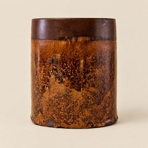 May include: A cylindrical ceramic vessel featuring a smooth, dark brown upper band that transitions into a textured, mottled glaze of amber, copper, and deep brown tones across the lower body, creating a distinct contrast in surface finish.