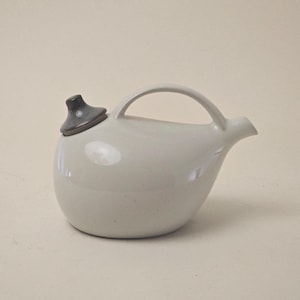 May include: A light grey ceramic teapot with a rounded body, a curved handle, and a spout. The lid is dark grey. The teapot is designed for brewing and serving tea.