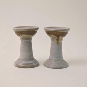 May include: Two ceramic candle holders. Each has a light gray, textured pedestal base and stem, topped with a brown-glazed bowl. The candle holders are displayed on a neutral background.
